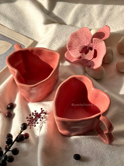 Two pink heart-shaped ceramic mugs on a light-colored surface, surrounded by decorative items including a pink orchid and small red berries.